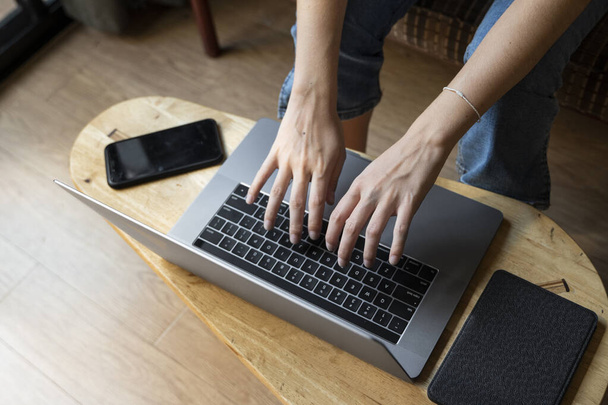 Vrouw zit op een bank en gebruikt een laptop aan houten tafel. Studeer en werk online, freelance. Zelfstandig meisje werkt met haar notebook zitten op een bank met een telefoon en ereader op tafel. - Foto, afbeelding