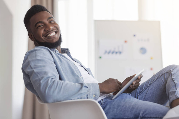 Sonriente chico afroamericano sentado en el trabajo con la tableta digital
 - Foto, imagen