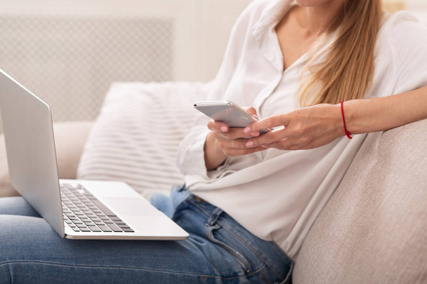 Неузнаваемая женщина с помощью телефона сидит с ноутбуком на диване в помещении
 - Фото, изображение