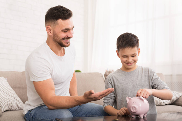 Vader leert zijn zoon geld te besparen, jongen stopt munten in spaarpot - Foto, afbeelding