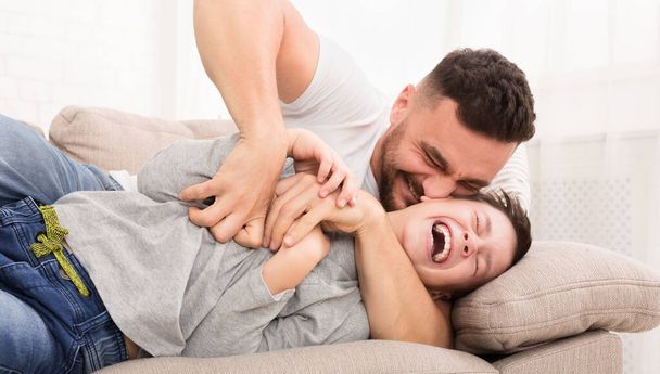 Playful dad tickling his laughing son, laughing on couch together - 写真・画像