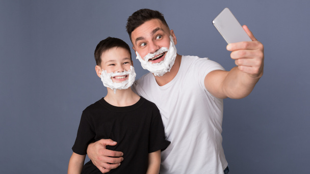 Счастливые папа и сын с пеной для бритья делают селфи по мобильному телефону
 - Фото, изображение