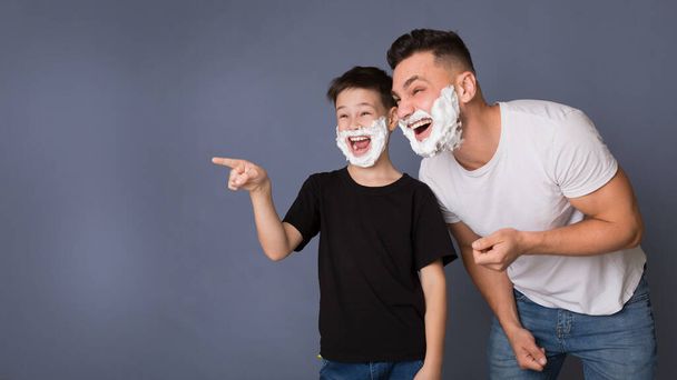 Emocionado padre e hijo con espuma de afeitar en las caras señalando a un lado
 - Foto, Imagen