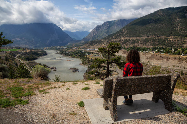 Girl Sitting on a Bench and enjoying the Beautiful View of a small Town, Lillooet, during a sunny and cloudy summer day. Located in the Interior British Columbia, Canada. - Фото, зображення