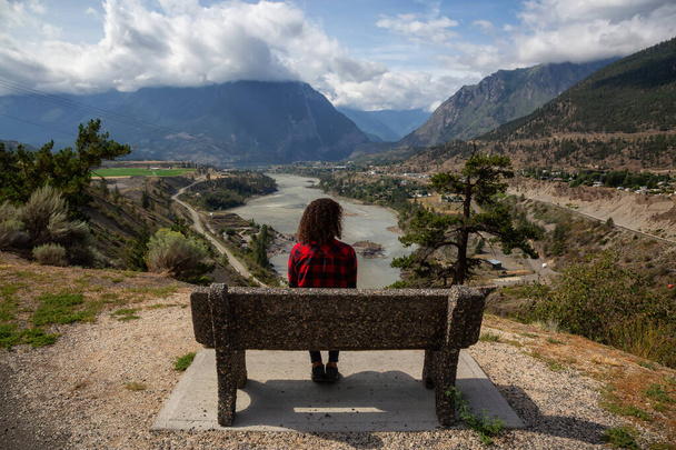 Girl Sitting on a Bench and enjoying the Beautiful View of a small Town, Lillooet, during a sunny and cloudy summer day. Located in the Interior British Columbia, Canada. - Photo, image