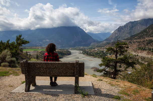 Girl Sitting on a Bench and enjoying the Beautiful View of a small Town, Lillooet, during a sunny and cloudy summer day. Located in the Interior British Columbia, Canada. - Foto, imagen