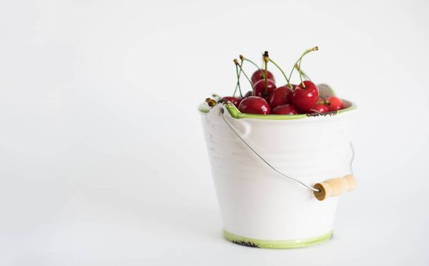 Ripe red cherry in white garden enamel bucket with a green border on a white background - Foto, imagen