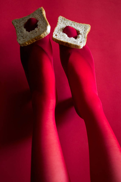 beautiful female legs in red tights hold slices of bread with their feet on a red background photo - Photo, Image
