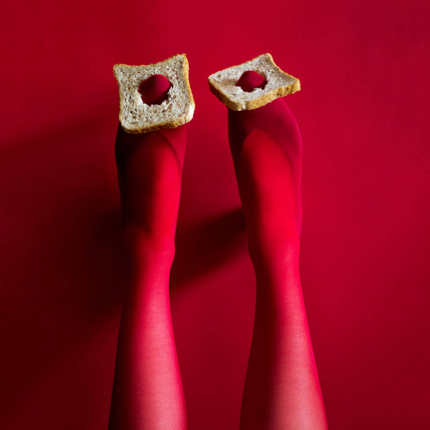 Foto schöne weibliche Beine in roten Strumpfhosen halten Toastscheiben auf dem Kopf auf rotem Hintergrund - Foto, Bild