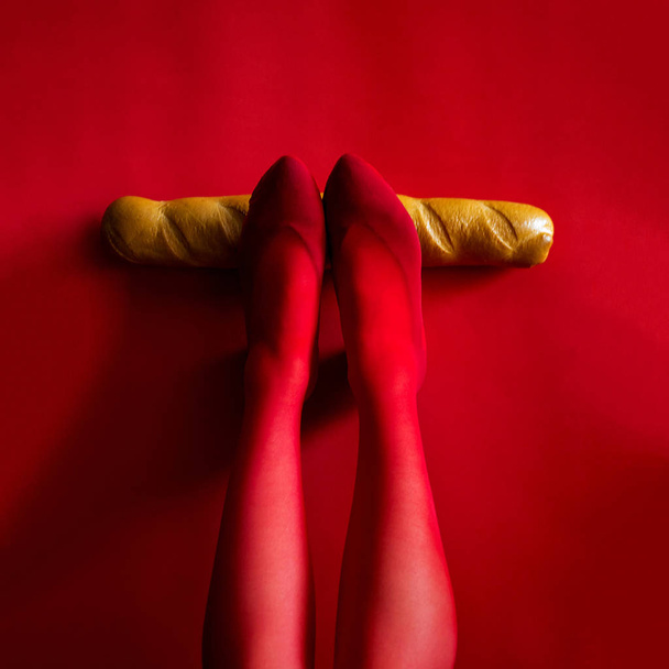 photo female legs in red tights hold a baguette horizontally on a red background - Photo, Image