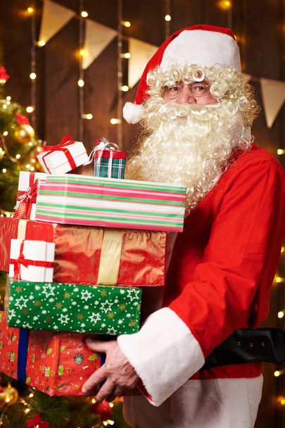 Père Noël posant avec de nombreux cadeaux, assis à l'intérieur près de l'arbre de Noël décoré avec des lumières Joyeux Noël et Joyeuses Fêtes
! - Photo, image