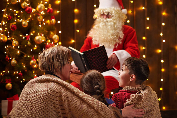 サンタクロースは家族のために本を読んでいます。照明付きの装飾されたクリスマスツリーの近くに屋内に座っている母親と子供-メリークリスマスとハッピーホリデー! - 写真・画像