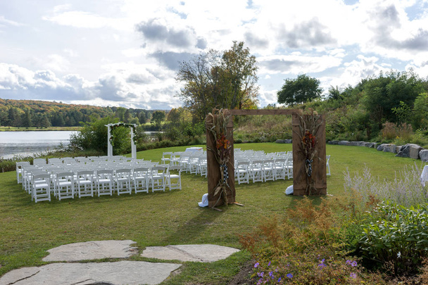 Miejsce ceremonii ślubnej w Lakeshore. Łuk ślubny ozdobiony kwiatami i białymi krzesłami z każdej strony łuku na zewnątrz - Zdjęcie, obraz