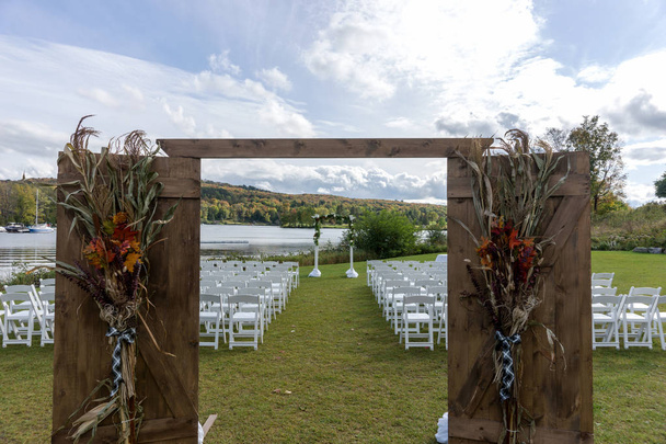 Place for wedding ceremony at the Lakeshore. Wedding arch decorated with flowers and white chairs on each side of archway outdoors - Photo, Image