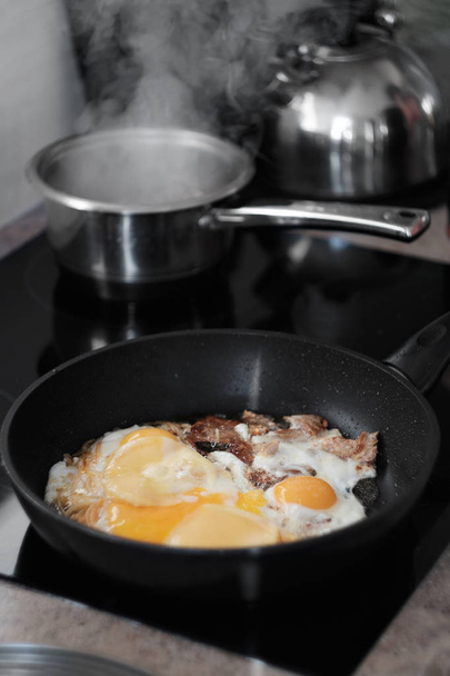 Los huevos fritos se cocinan en una sartén, el ganado está hirviendo agua en la parte posterior
 - Foto, Imagen