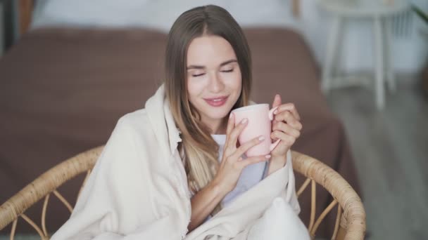 Portrait of smiling woman wrapped in plaid drinks hot tea, looks at camera - Materiał filmowy, wideo