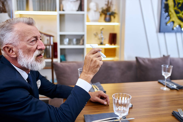 Portrait senior man grey haired sit in restaurant looking electronic cigarette - Φωτογραφία, εικόνα