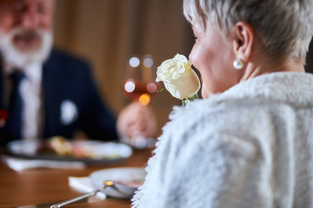 Elegant senior couple dressed up in tuxedo and white clothes, woman holding a Valentines Day rose. - Фото, изображение