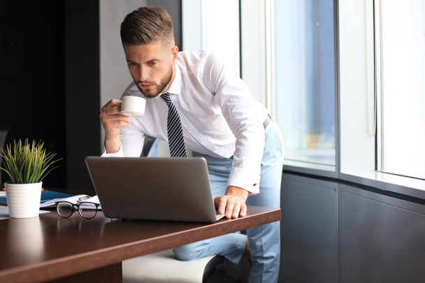 Вдумчивый молодой бизнесмен пользуется компьютером и пьет кофе во время работы в офисе
 - Фото, изображение