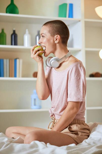 Beautiful caucasian girl sits in white bed early in morning, eats green apple - Foto, imagen