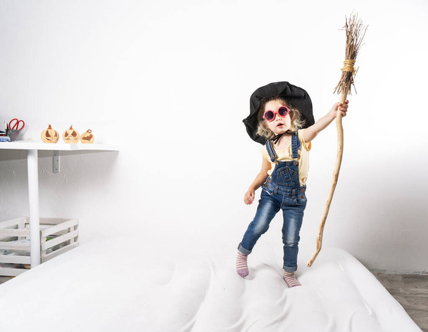 Little girl in a witch hat jumps on a mattress with a broom. - Fotó, kép