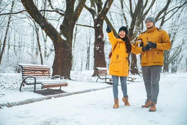пара прогуливаясь по снежному городскому парку пьет кофе, чтобы показать что-то прямо
 - Фото, изображение