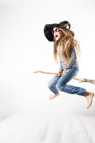 Schoolgirl in a witch hat jumps on a mattress and flies on a broomstick. Childrens lifestyle portrait. - Photo, image