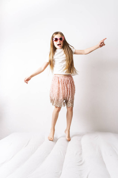A little girl in pink sunglasses and a pink skirt jumps on a white mattress. - Fotó, kép