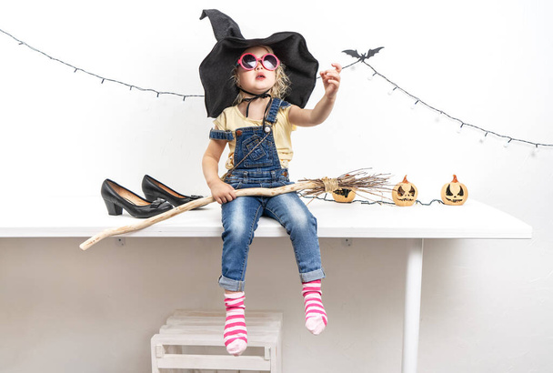 Happy Halloween: A little girl sits on a table in a witch hat with a broom. - Fotó, kép