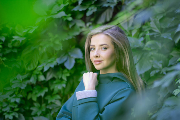 Portrait of a beautiful girl in a green coat on a background of ivy . - Zdjęcie, obraz