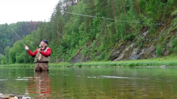 Pesca con mosca. Un pescador experimentado en ropa roja está parado en el agua. Río de montaña. Un hombre con barba, parecido a Papá Noel. Camping, pesca deportiva
. - Imágenes, Vídeo