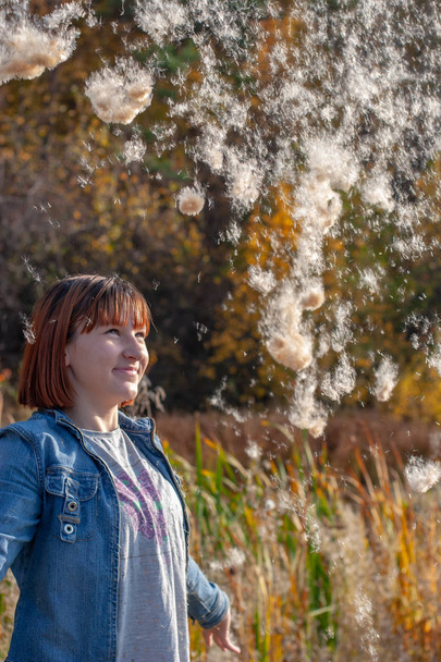 A teenage girl with red hair smiling throws up fluff from a cattail and he beautifully falls down. Airy fluff like parachutes. Autumn forest and grass on a blurred background. - Photo, Image
