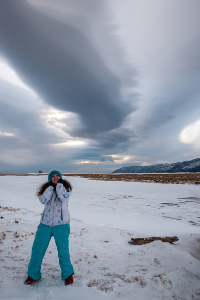 Κορίτσι τραβώντας ένα καπέλο το χειμώνα κοιτάζει ενδιαφέροντα σύννεφα στον ουρανό. Κορίτσι με μακριά μαύρα μαλλιά με χειμωνιάτικα ρούχα. Κάθετο πλαίσιο. - Φωτογραφία, εικόνα