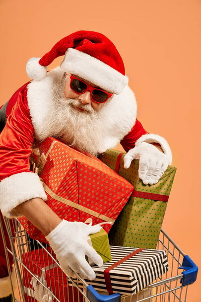 Homem envelhecido em traje de Santa com caixas de presente no carrinho de compras
 - Foto, Imagem