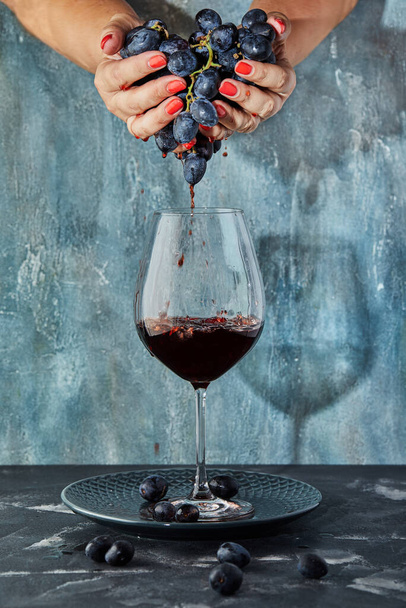 Kobiece dłonie wyciskające winogrona do kieliszka z winem, na niebieskim tle. Koncepcja produkcji wina - Zdjęcie, obraz