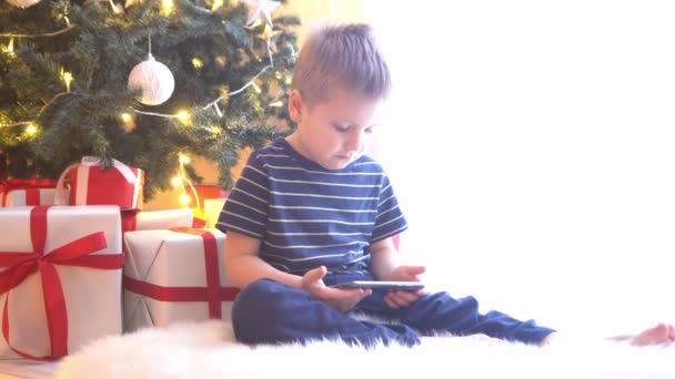 Attractive little boy watching cartoons in front of Christmas tree with balls and lights. Kid preparing for the New Year at home. - Footage, Video