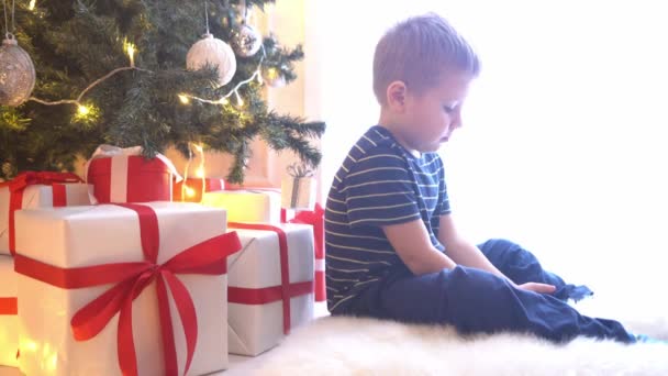 Attractive little boy watching cartoons in front of Christmas tree with balls and lights. Kid preparing for the New Year at home. - 映像、動画