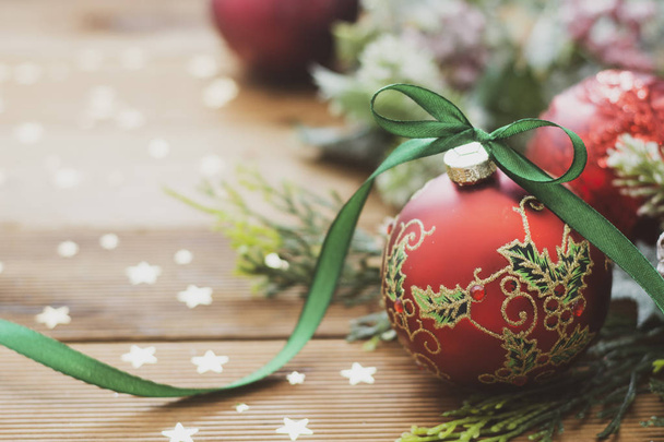 Christmas background, greeting card. Red beautiful bauble with green ribbon, fir branches, winter decoration, gift boxes over wooden background. Copy space. - 写真・画像