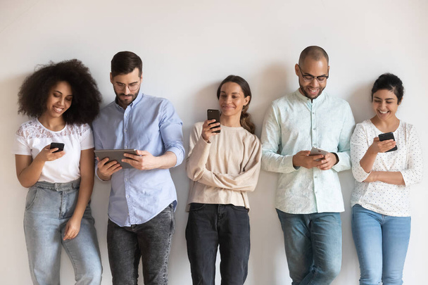 Smiling diverse people using phones, standing in row near wall - Photo, Image