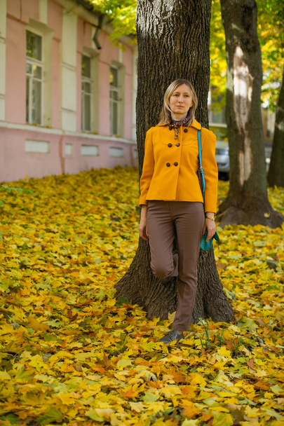 Blondynka w żółtej kurtce pozuje na jesiennej ulicy z czerwono-żółtymi liśćmi. - Zdjęcie, obraz
