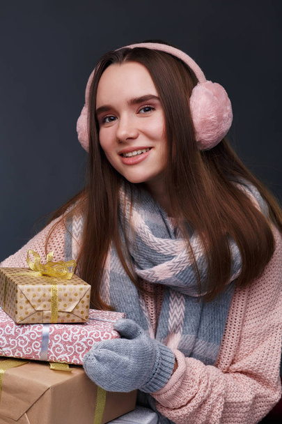 Young beautiful smiling girl in knitting pullover and pink fluffy earmuffs holding gift boxes on a gray background. Xmas model with long straight hair. Winter holidays, Christmas, New Year concept.  - Fotoğraf, Görsel