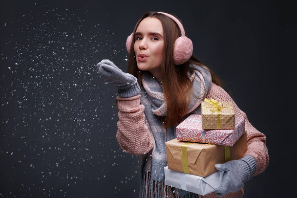 Fiatal, gyönyörű, mosolygós lány kötött pulóverben és rózsaszín, bolyhos fülvédővel, kezében ajándékdobozokkal és fehér hópelyhekkel, szürke háttérrel. Téli ünnepek, Karácsony, Szilveszter koncepció.  - Fotó, kép