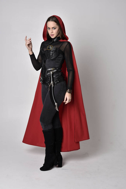 full length portret van een mooie brunette vrouw in zwart lederen fantasie kostuum met lange rode superheld cape. staande houding op een studio achtergrond. - Foto, afbeelding