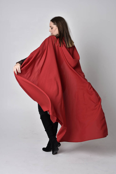 Ganzkörperporträt einer hübschen brünetten Frau in schwarzem Leder-Fantasiekostüm mit langem roten Superhelden-Umhang. stehende Pose auf einem Studiohintergrund. - Foto, Bild