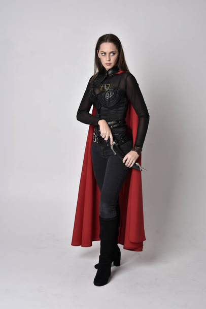 retrato de cuerpo entero de una guapa morena vestida con un disfraz de fantasía de cuero negro con capa larga de superhéroe rojo. pose de pie sobre un fondo de estudio
. - Foto, imagen