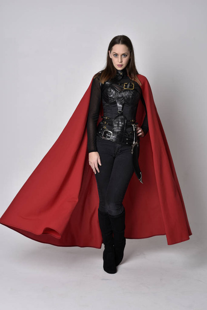 Uzun boylu, esmer, siyah deri fantezi kostümlü, uzun kırmızı süper kahraman pelerinli bir kadının portresi. Stüdyo arka planında ayakta durma. - Fotoğraf, Görsel
