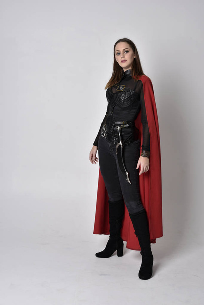 full length portrait of a pretty brunette woman wearing black leather fantasy costume with long red superhero cape. standing pose on a studio background. - Photo, Image