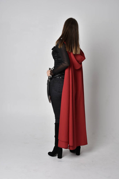 Ganzkörperporträt einer hübschen brünetten Frau in schwarzem Leder-Fantasiekostüm mit langem roten Superhelden-Umhang. stehende Pose auf einem Studiohintergrund. - Foto, Bild