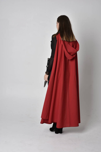 長い赤いスーパーヒーローのケープと黒革のファンタジーの衣装を身に着けているかなりブルネットの女性の完全な長さの肖像画。スタジオの背景に立ちポーズをとって. - 写真・画像