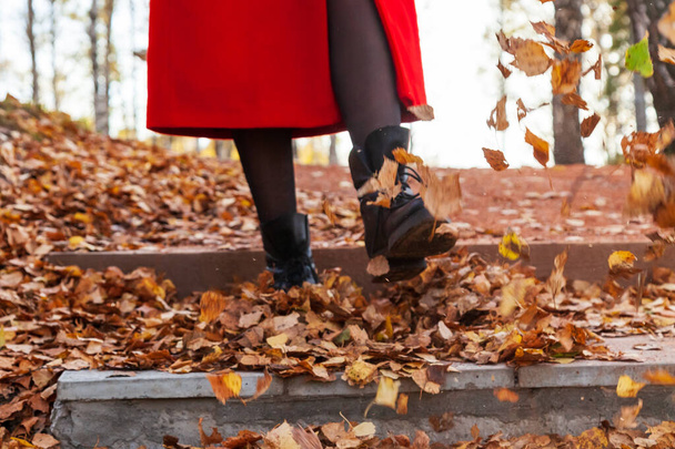 Девушка в чёрных сапогах и красном пальто пинает жёлтую и красную листву.
 - Фото, изображение